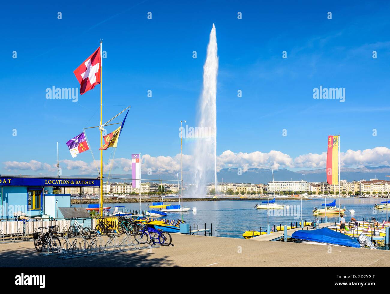 Die Schweizer Flagge fliegt an einem sonnigen Morgen am Ufer des Genfersees in Genf, Schweiz, mit dem Wasserstrahlbrunnen in der Ferne. Stockfoto