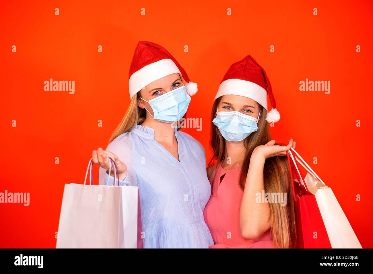 Weibliche Freunde tragen chirurgische Maske und Weihnachtsmütze halten einkaufen Taschen auf einem leuchtend roten orangen Hintergrund Stockfoto