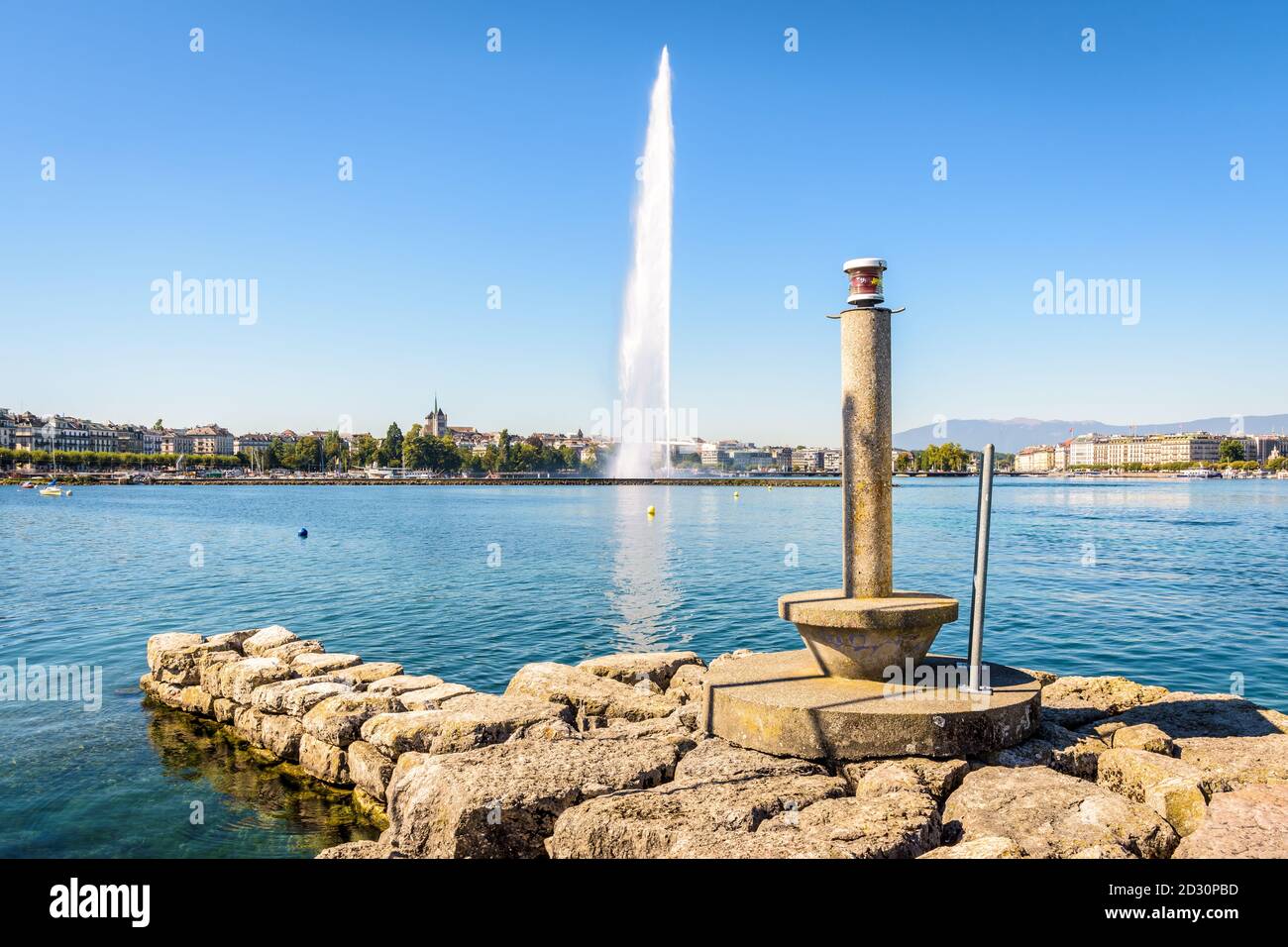 Die Stadt und die Bucht von Genf, Schweiz, mit ihrem Wasserstrahlbrunnen auf dem Genfer See, von einem Steg mit einem Leuchtfeuer an einem sonnigen Sommermorgen gesehen. Stockfoto