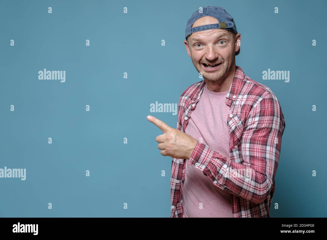 Lustiger Mann zeigt Zeigefinger zur Seite und schaut mit einem seltsamen Gesichtsausdruck auf die Kamera. Speicherplatz kopieren. Blauer Hintergrund. Stockfoto