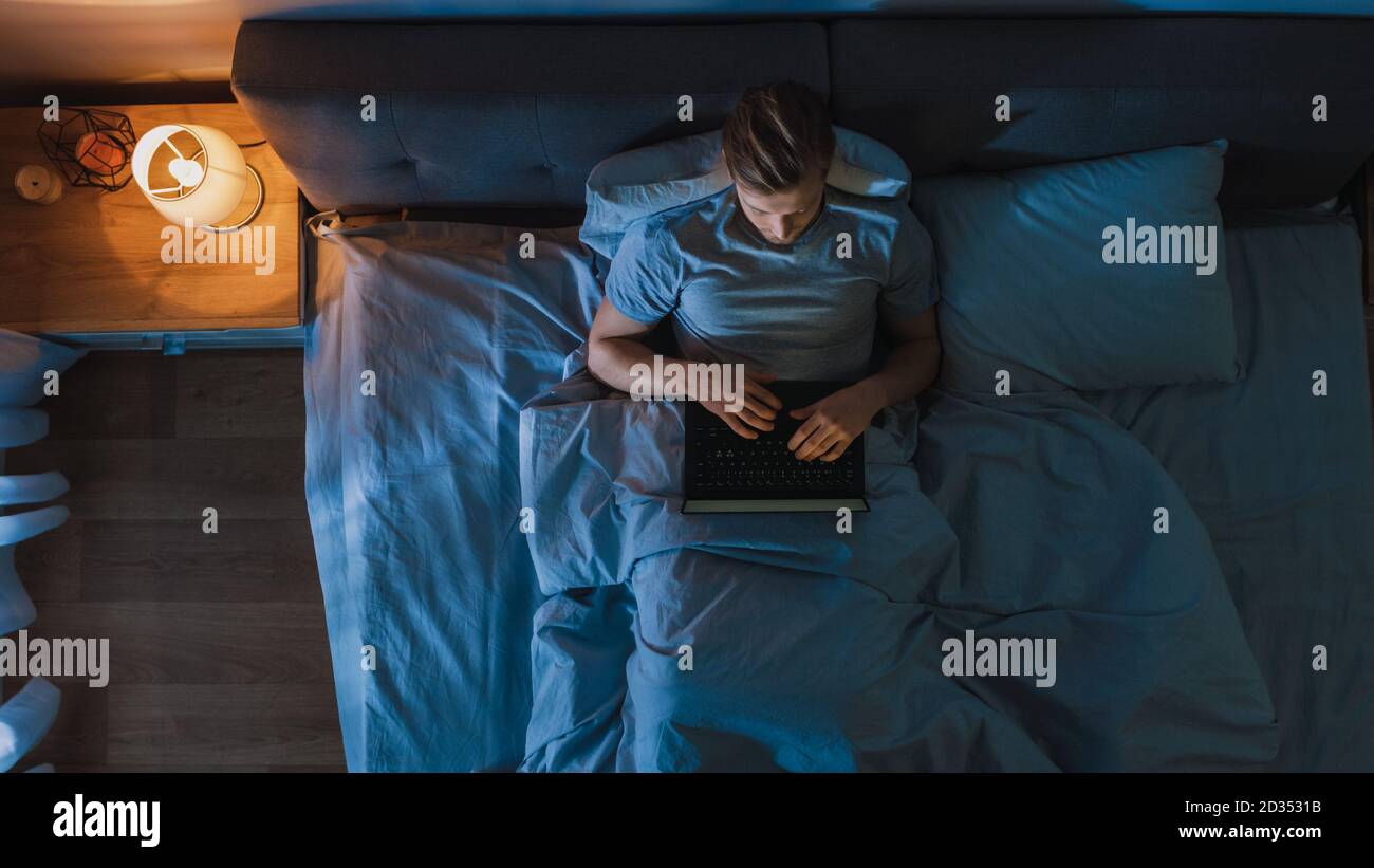 Blick von oben junger Mann im Bett, der nachts an einem Laptop-Computer arbeitet. Schüler, die sich auf Prüfungen vorbereiten, außergewöhnlich engagierter Projektmanager, Abschluss Stockfoto