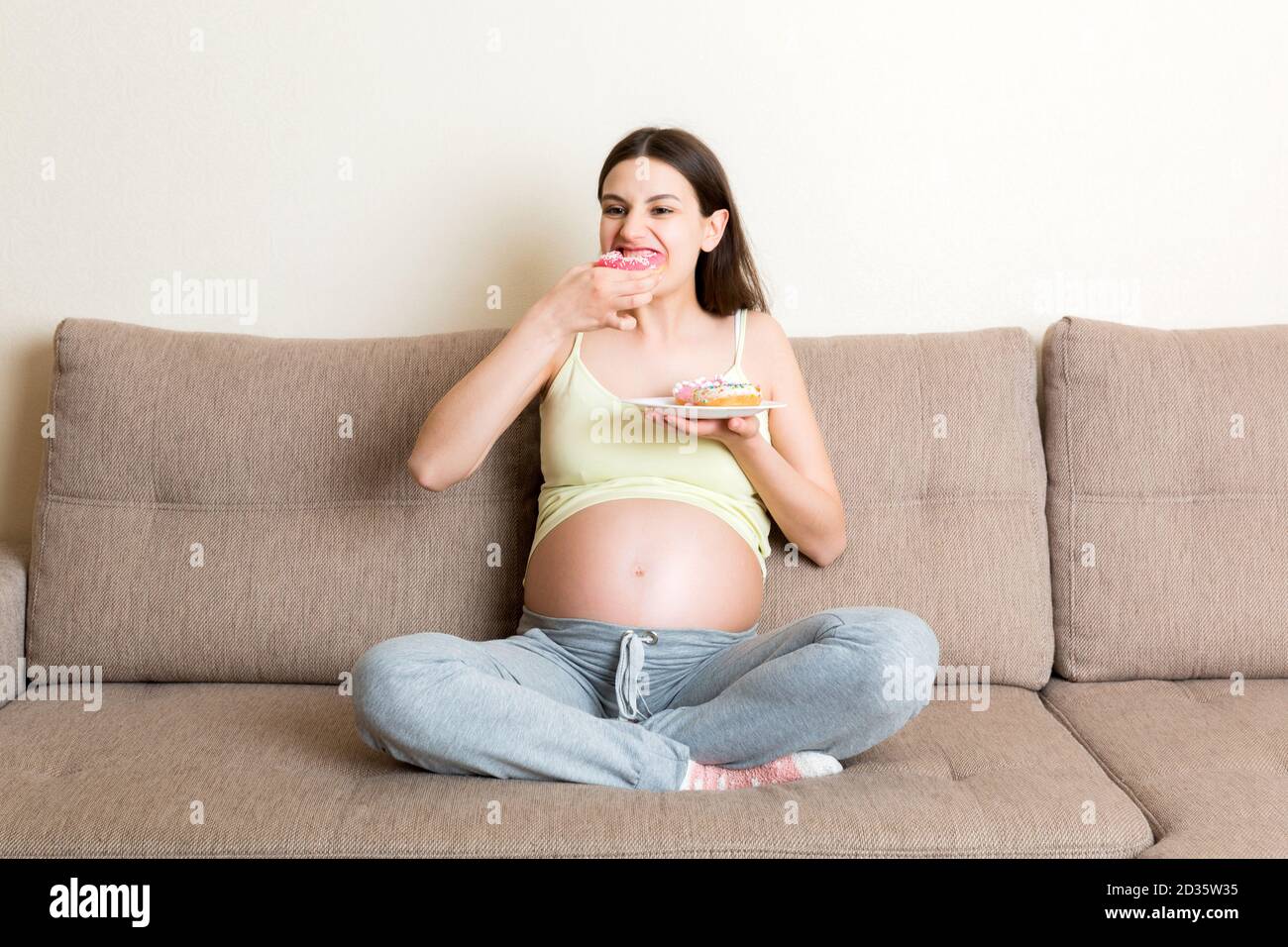 Schwangere Frau genießt es, verschiedene Donuts auf dem Sofa ruhen zu essen. Ungesunde Desserts während der Schwangerschaft Konzept. Stockfoto