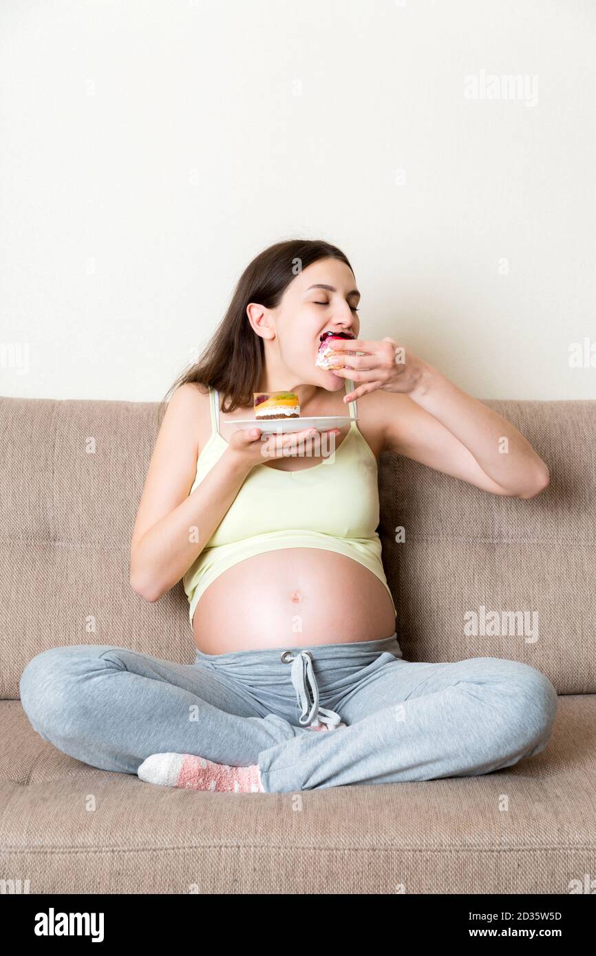 Hungrige schwangere Frau essen Gebäck mit großer Freude auf dem Sofa zu Hause entspannen. Happy Schwangerschaft Konzept. Stockfoto