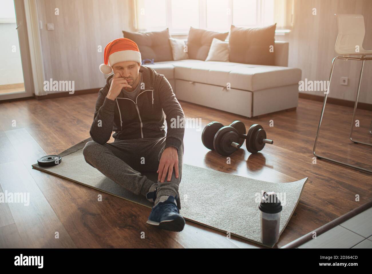 Der junge gewöhnliche Mann geht zu Hause zum Sport. Traurig, aufgeregt und gelangweilt Kerl in roten Hut sitzen auf Matte nach dem ersten Training für sich. Amateur und nicht Stockfoto