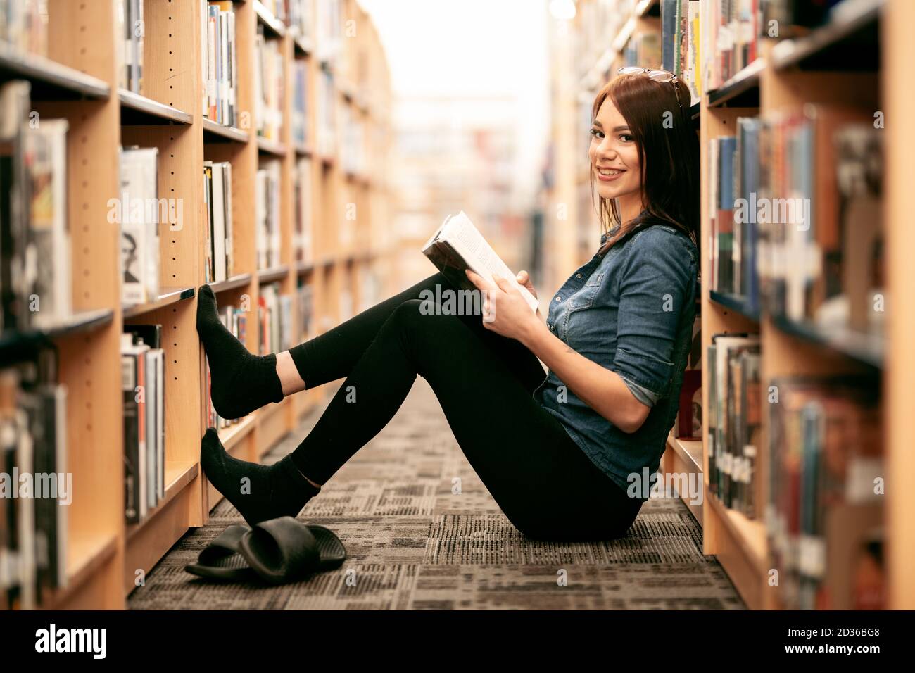 Eine einsame junge Frau, die in ihrer örtlichen Bibliothek Bücher liest, mit Blick auf Bücherregale und Bücherregale. Stockfoto