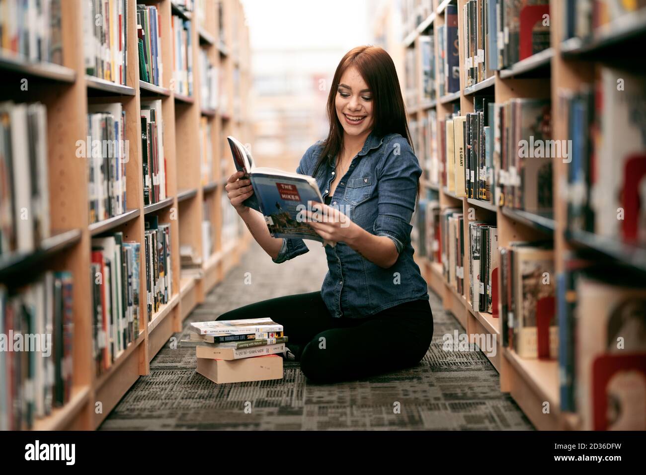 Eine einsame junge Frau, die in ihrer örtlichen Bibliothek Bücher liest, mit Blick auf Bücherregale und Bücherregale. Stockfoto