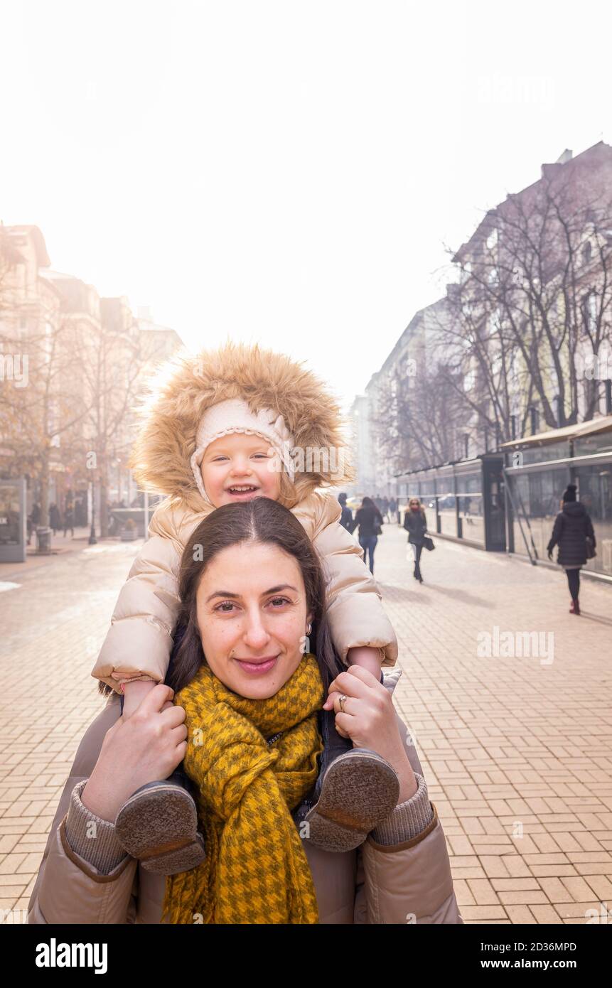 Glückliche Mutter trägt Tochter auf ihren Schultern in Outdoor-Stadt Straßenszene im Winter, Vitosha Boulevard, Sofia, Bulgarien Stockfoto