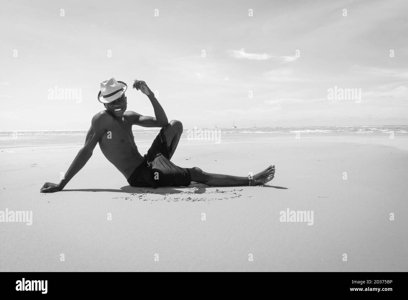 Schwarzer junger madagassischer Mann mit Hut und großem Lächeln, der am weißen Sandstrand vor dem Meer in einer sehr klassischen Position sitzt und sich an die Hand lehnt Stockfoto