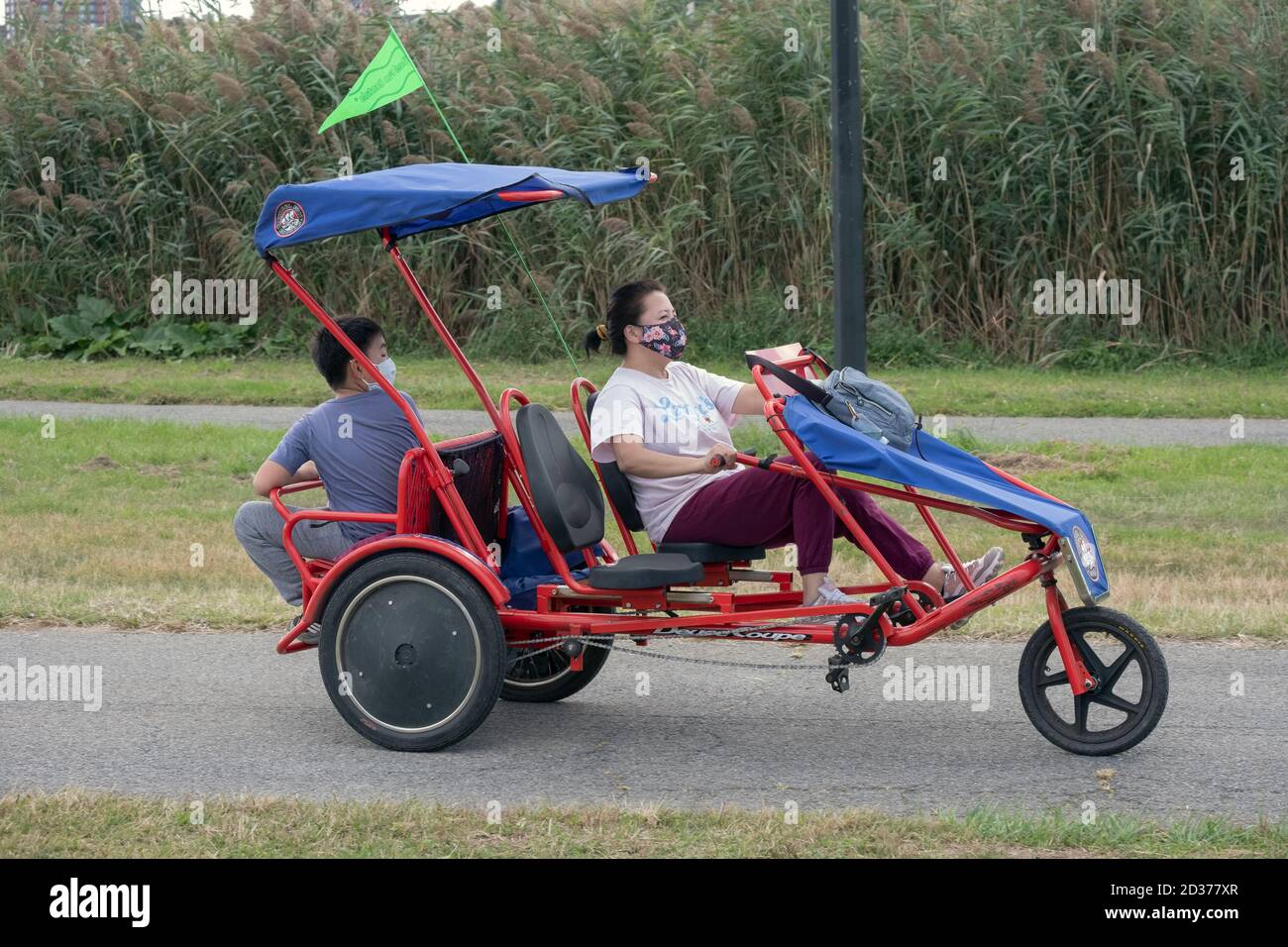 Eine asiatisch-amerikanische Mutter und Sohn mit Masken auf einem Rad Spaß 3 Rad surrey Zyklus in Flushing Meadows Corona Park in Queens, New York City. Stockfoto