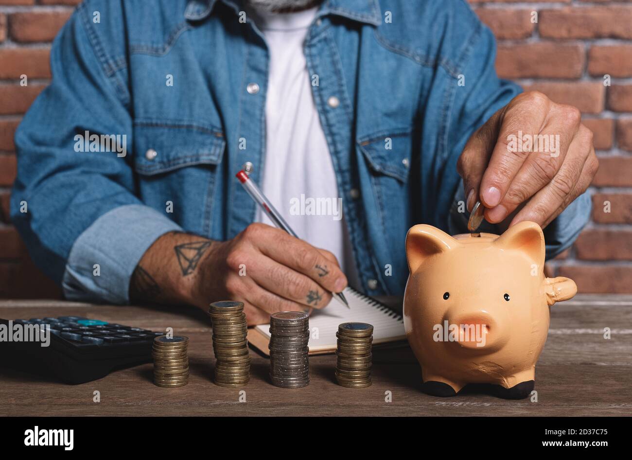 Hipster Mann Putting Münze in Schwein Sparschwein mit Haufen von Münzen auf dem Tisch, um Geld zu sparen. Stockfoto