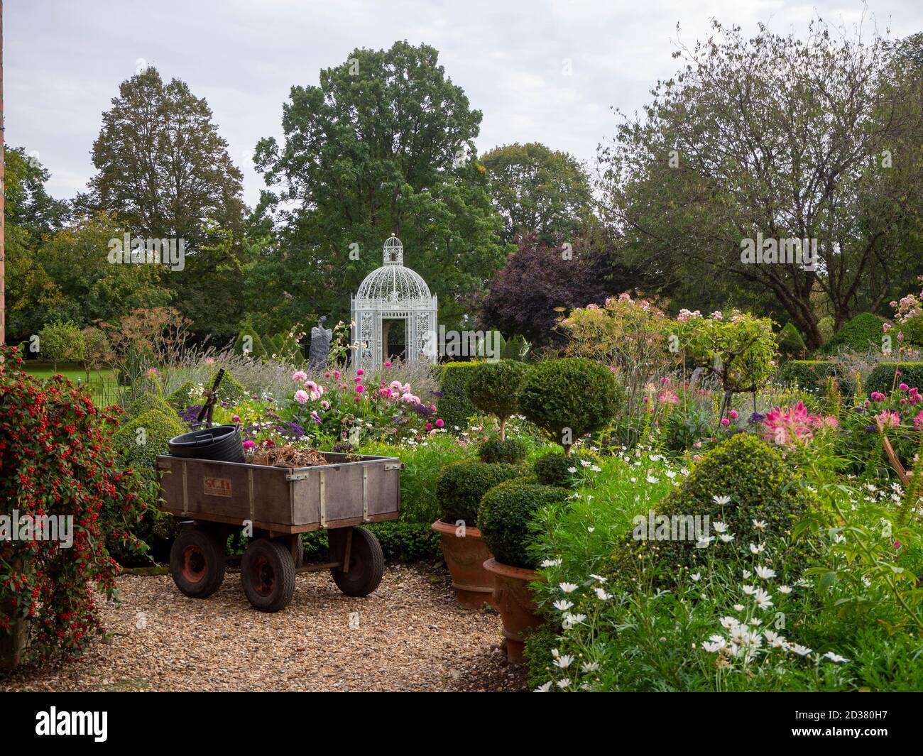 Chenies Manor Garten. Ein Handwagen ruht nach der Arbeit im Garten auf dem Weg. Ein Vogelkäfigpavillon im Parterre hinter den rosa und weißen Blüten. Stockfoto