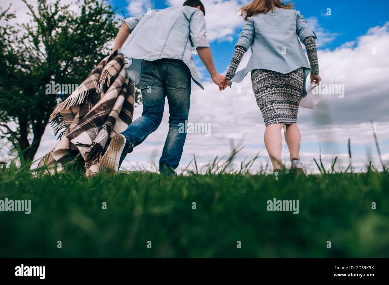 Junges Paar in der Liebe mit einem karierten karierten kariert läuft über ein grünes Feld halten Hände auf einem Hintergrund von blauem Himmel. Rückansicht und Unteransicht. Getöntes Bild Stockfoto