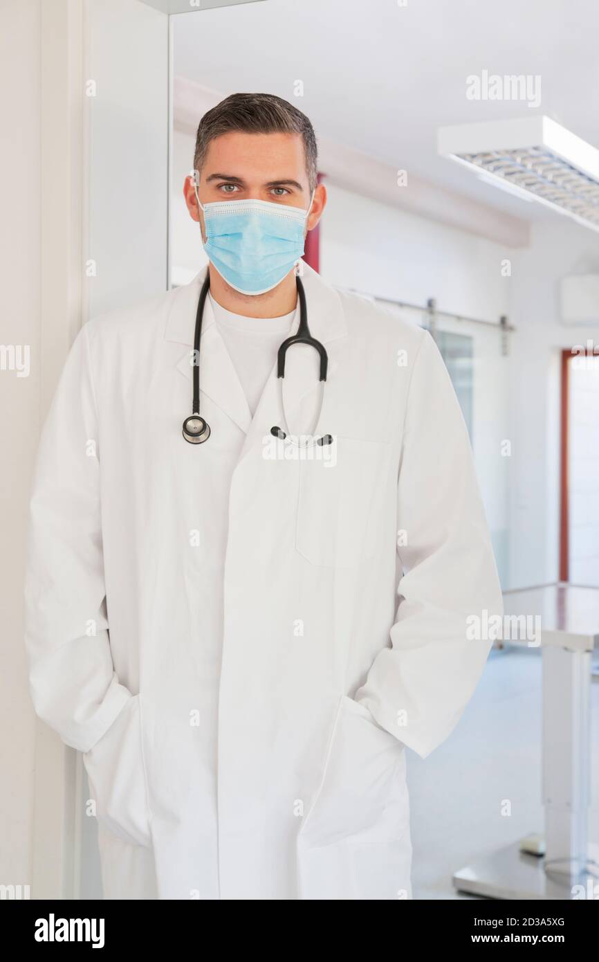 Junger selbstbewusster Arzt oder Chirurg mit medizinischer Maske im Stehen Vorderseite seiner Operation im verschwommenen Hintergrund Stockfoto