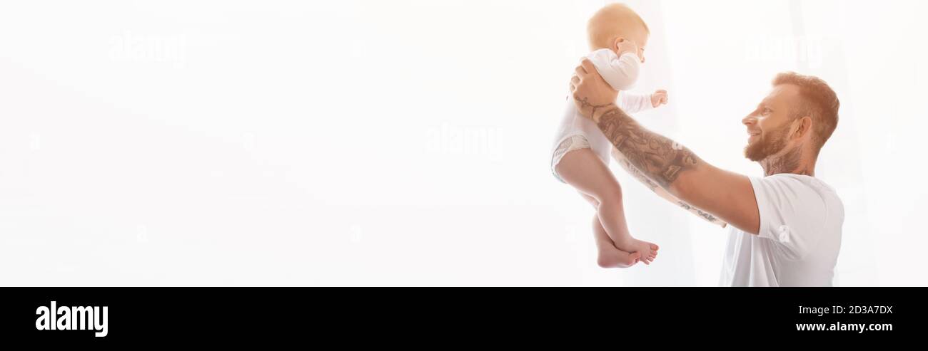 Seitenansicht des jungen tätowierten Mannes, der Säugling Sohn in ausgestreckten Händen hält, Website-Header Stockfoto