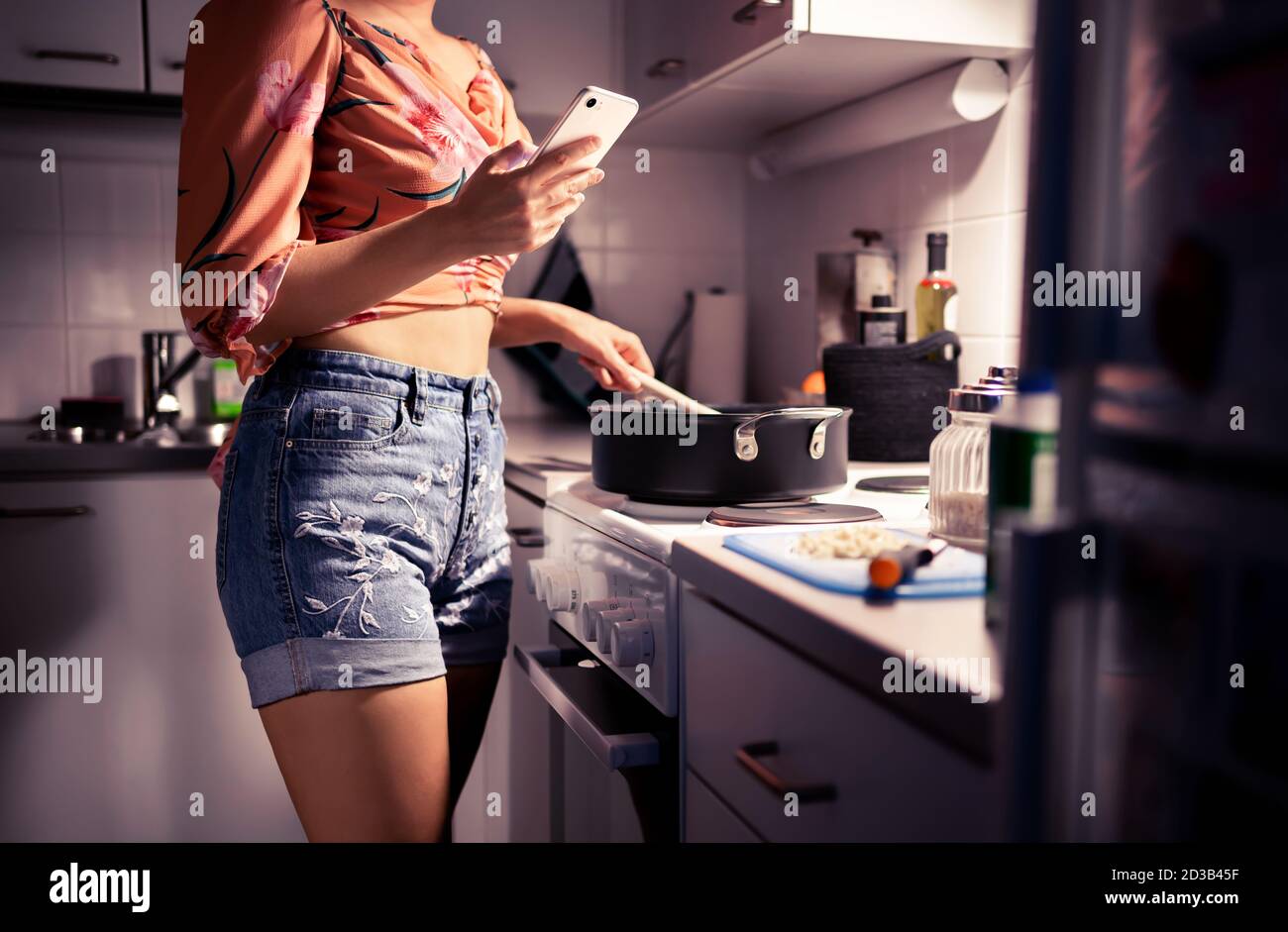 Junge Frau Kochen und mit Telefon. Person, die ein Abendessen vorbereitet und mit dem Smartphone SMS schreibt. Ernährung oder eine gesunde Lebensweise. Wohnküche in der Nacht. Stockfoto