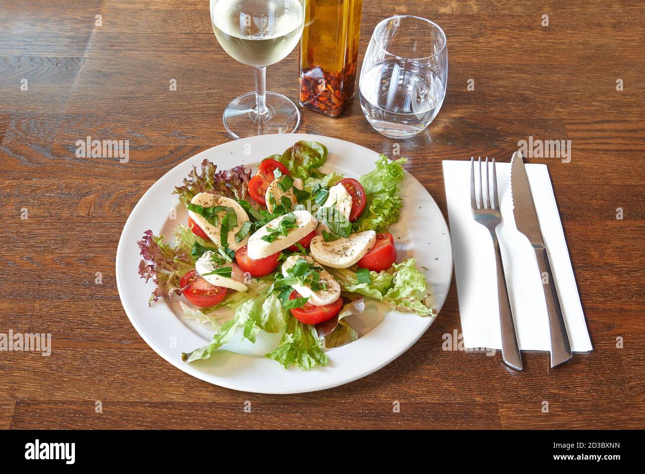 Besteck auf braunem Holztisch und weißer Teller mit Tomaten und Mozzarella auf Salat. Stockfoto