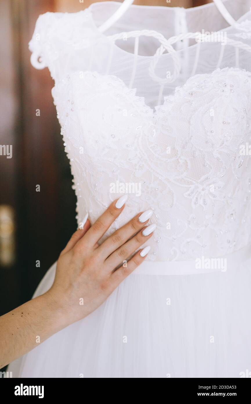 Nahaufnahme der Hand der Braut berührt eine schöne weiße Hochzeitskleid, das auf einem weißen Kleiderbügel hängt. Bester Tag der Braut. Hochzeitsaccessoires. Stockfoto