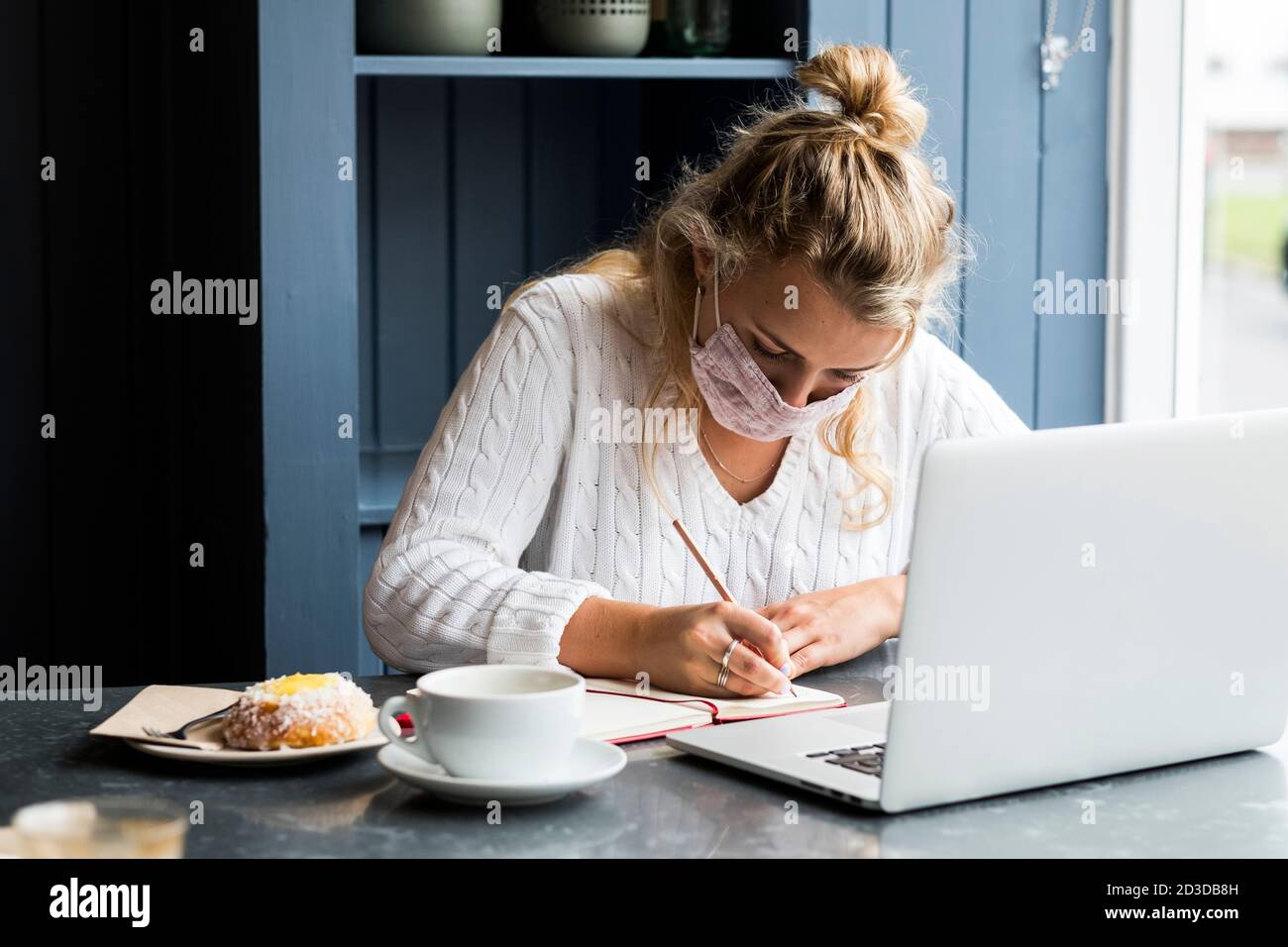 Junge blonde Frau trägt Gesichtsmaske allein sitzen an einem Café-Tisch mit einem Laptop-Computer, schriftlich in Notizbuch, arbeiten aus der Ferne. Stockfoto