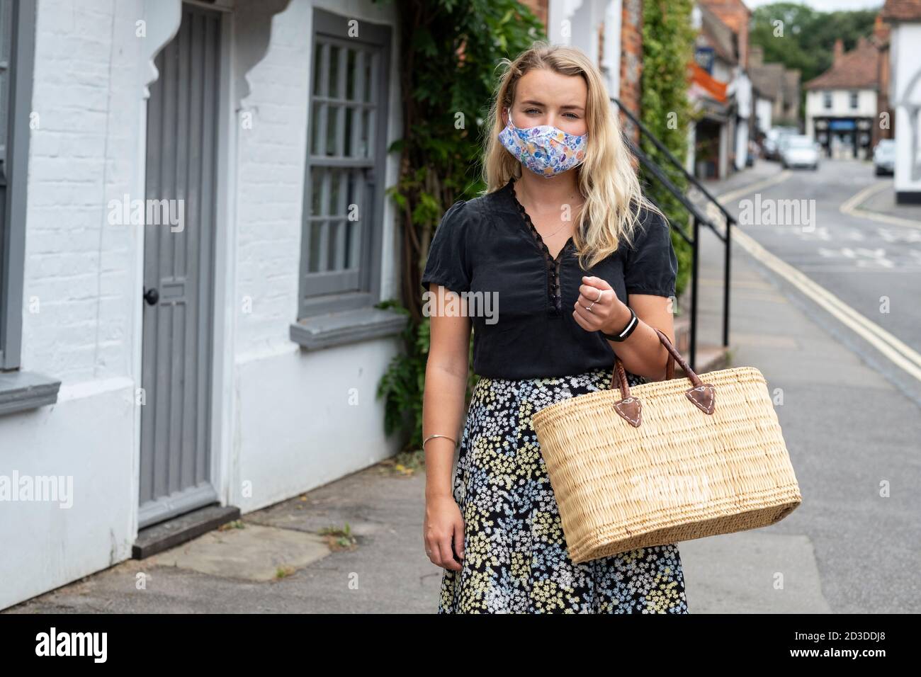 Junge blonde Frau trägt Gesichtsmaske zu Fuß durch Dorf, mit Einkaufstasche. Stockfoto
