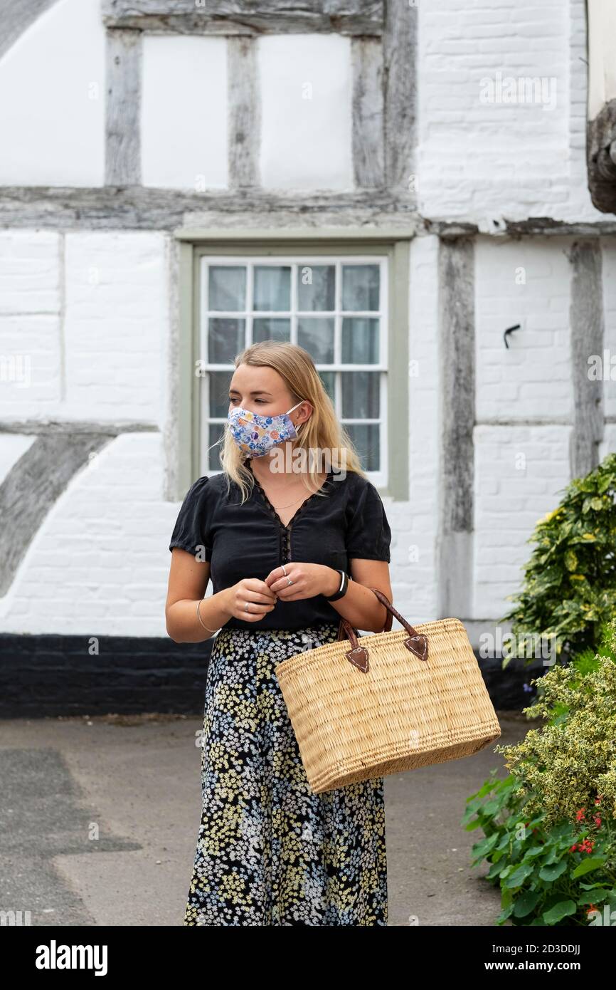 Junge blonde Frau trägt Gesichtsmaske zu Fuß durch Dorf, mit Einkaufstasche. Stockfoto