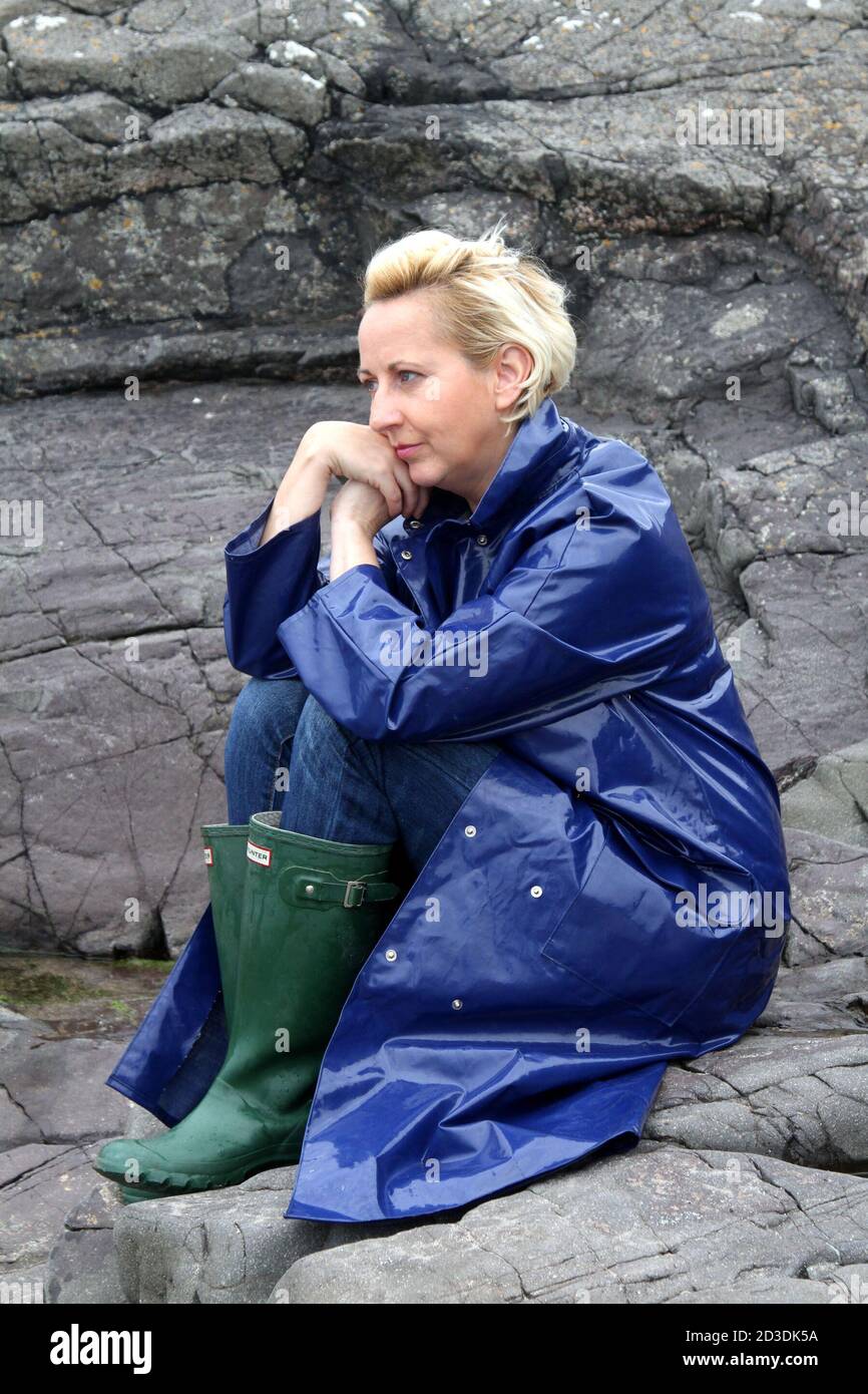 Weiß kaukasisch gefärbte blonde Frau mittleren Alters in ihren 40er Jahren  trägt einen blauen glänzenden PVC-Regenmantel aus einem Second-Hand-Shop  gekauft. Ich saß auf Felsen und sah nachdenklich aus Stockfotografie - Alamy