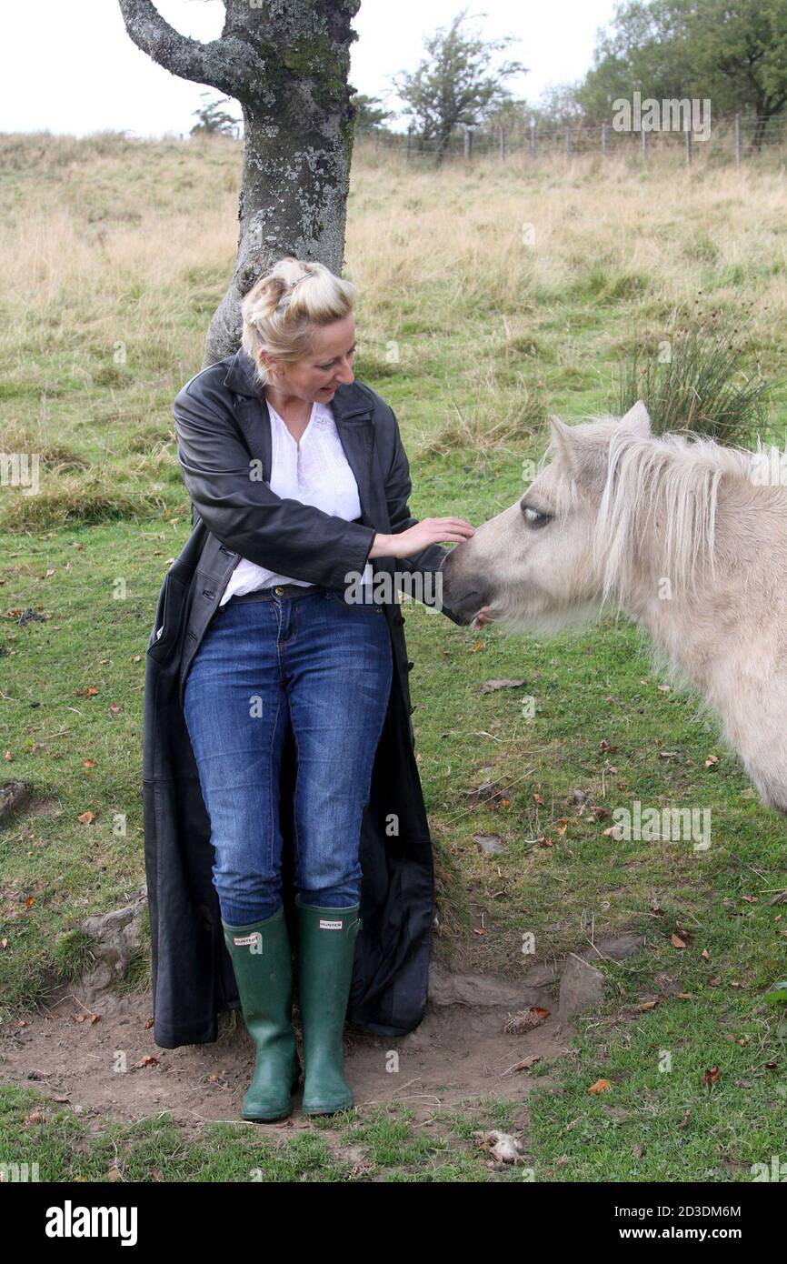 Weiß kaukasisch gefärbte blonde Frau mittleren Alters in ihren 40er Jahren trägt einen langen Ledermantel aus einem Second-Hand-Shop im Feld mit Pferden und Ponys gekauft. Stockfoto