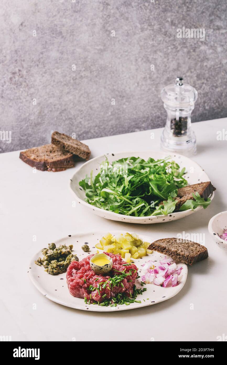 Beef Tartare mit Wachtelei, in der Schale, Roggenbrot, schneiden Gewürzgurken, Kapern, Zwiebeln, Schnittlauch und Rucola Salat in beschmutzt Keramik plat Stockfoto