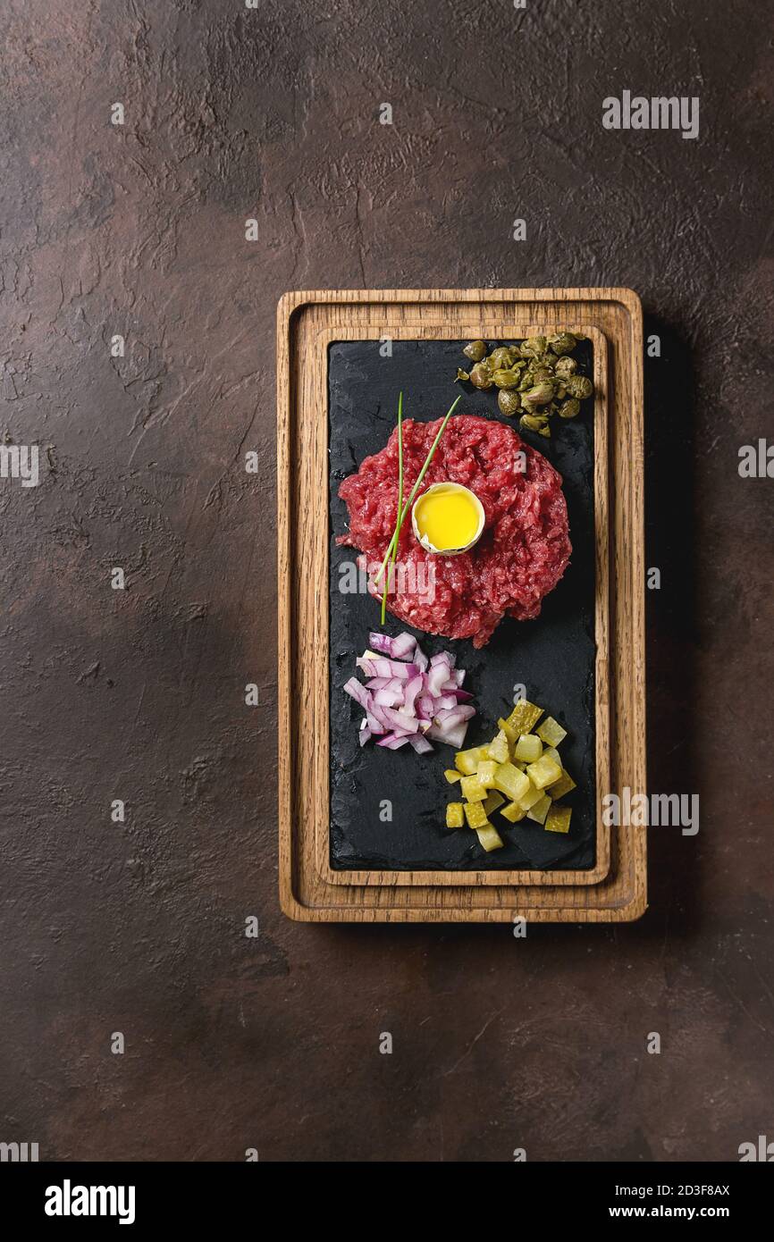 Beef Tartare mit Wachteleier in der Schale, schneiden Gewürzgurken, Kapern, Zwiebeln, Schnittlauch auf hölzernen schwarzer Schiefer mit Pfeffer Mühle auf dunklen serviert. Stockfoto