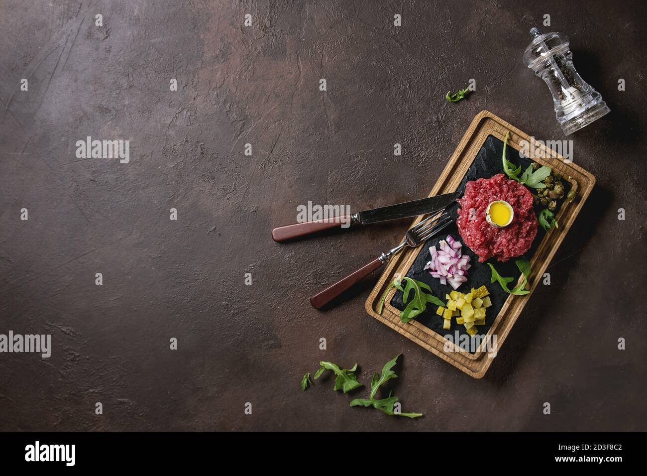 Beef Tartare mit Wachteleier in der Schale, schneiden Gewürzgurken, Kapern, rote Zwiebel, Schnittlauch, Rucola auf hölzernen schwarzer Schiefer mit Besteck auf serviert. Stockfoto
