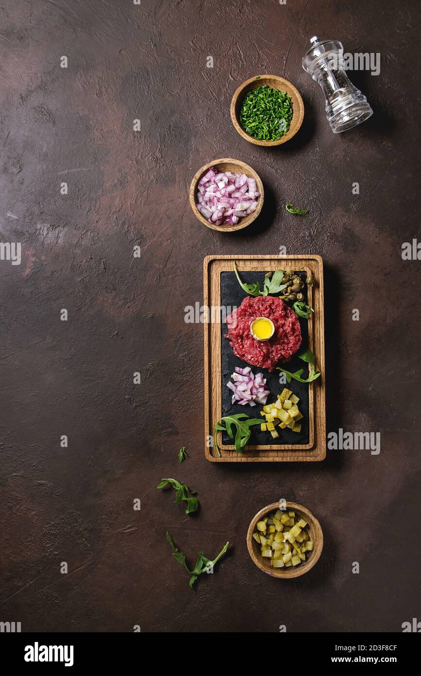 Beef Tartare mit Wachteleier in der Schale, schneiden Gewürzgurken, Kapern, rote Zwiebel, Schnittlauch, Rucola auf hölzernen schwarzer Schiefer mit Pfeffer mil serviert. Stockfoto