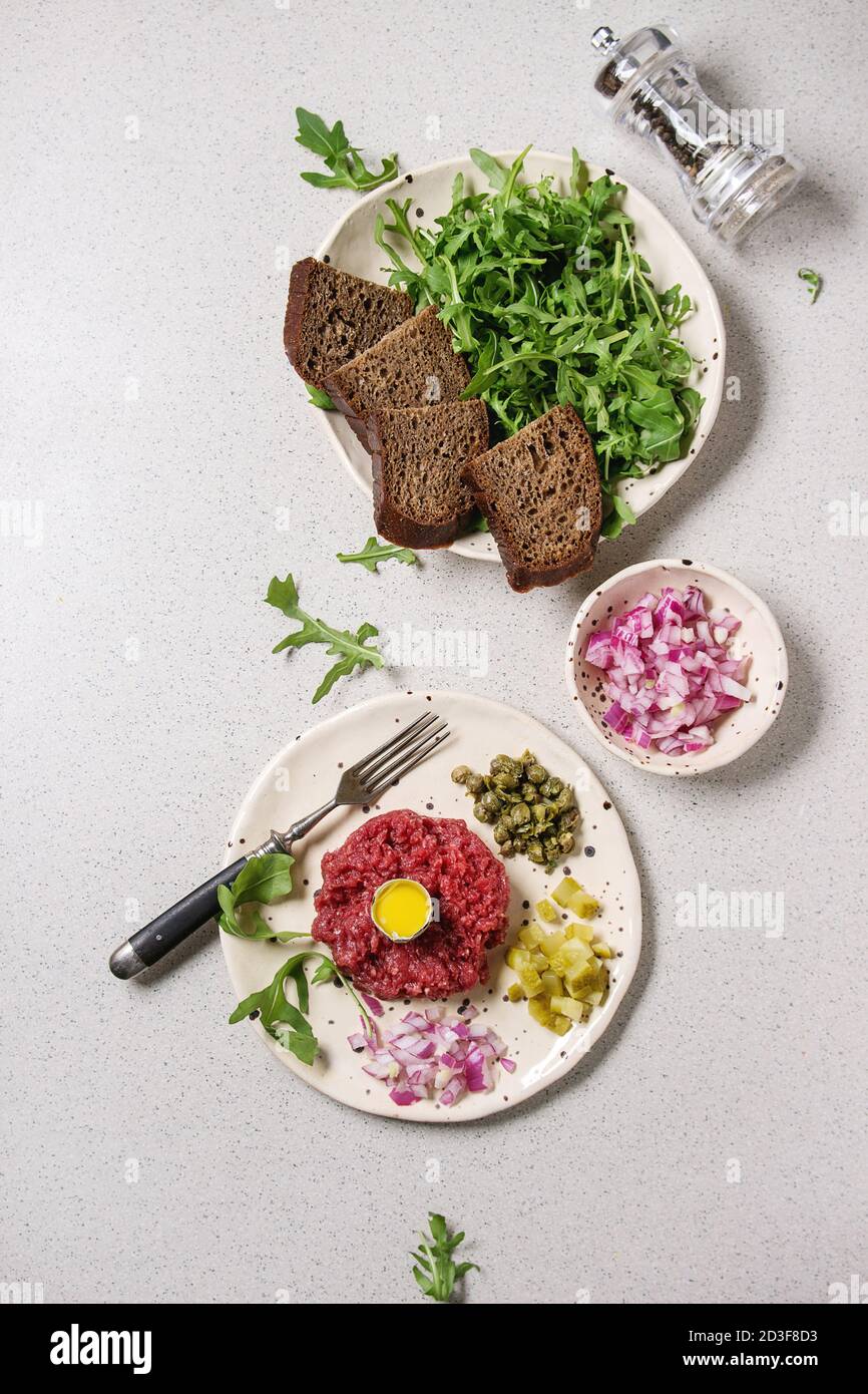 Beef Tartare mit Wachtelei, in der Schale, Brot, schneiden Gewürzgurken, Kapern, roten Zwiebeln, Rucola Salat in beschmutzt, keramischen Platten mit Pfeffer Stockfoto