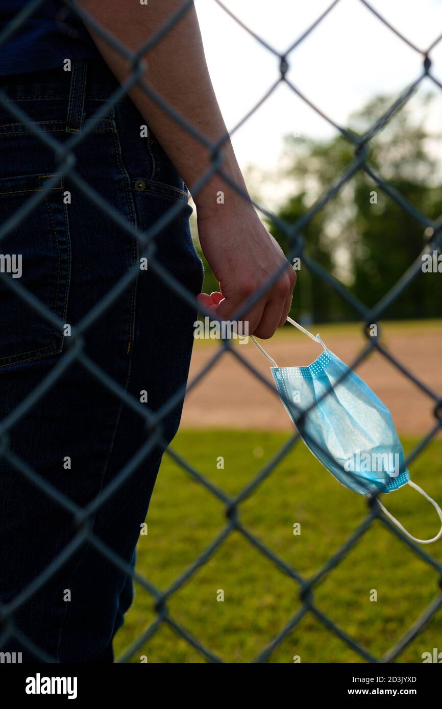 Blick durch den Kettengliederzaun am Baseballfeld auf einen jungen Mann mit einer medizinischen Maske, hinten beleuchtet. Stockfoto