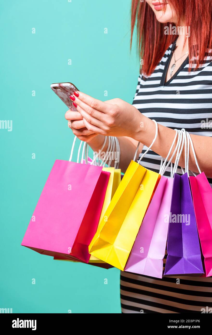 Frau, die Einkaufstaschen an den Händen hält und ein Smartphone benutzt. Nahaufnahme einer rothaarigen Frau, die mit bunten Papiertüten einkaufen. Stockfoto