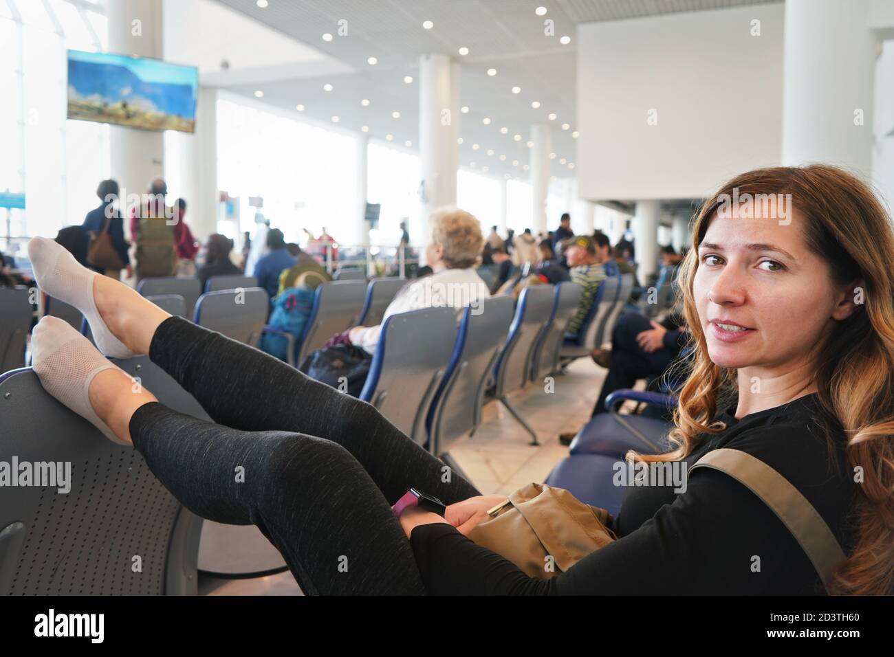 Junge Frau, die in der Flughafenhalle sitzt und müde aussieht, nachdem sie ein paar Stunden auf den Anschlussflug am frühen Morgen gewartet hat, die Beine auf dem Sitz vor dem h Stockfoto