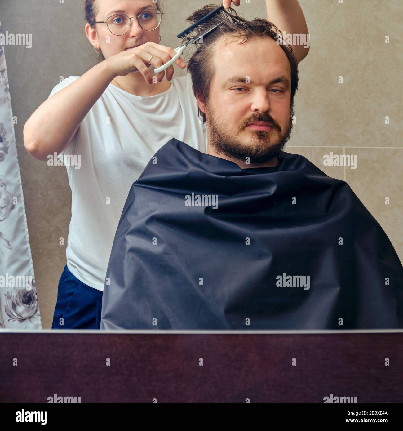 Frau schneidet einem Mann die Haare mit einer Schere vor dem Spiegel, Platz kopieren. Das Konzept der ungepflegten Erscheinung und Probleme in Isolation von der Coronavi Stockfoto