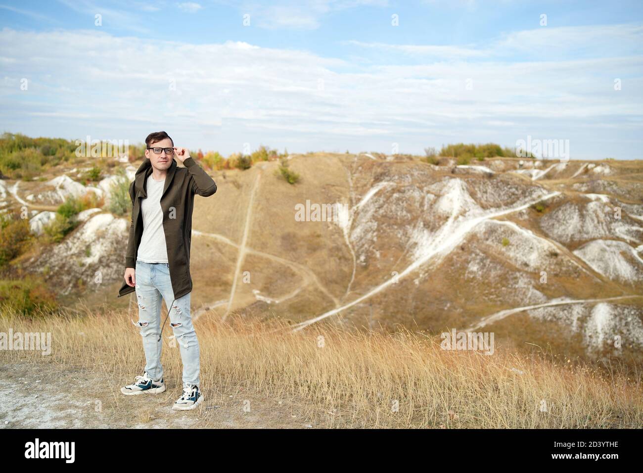 Mann mit Brille in Jeans, T-Shirt und Cape steht in hügeligem Gelände. Erwachsene Männchen in Kapuze passt seine Brille und genießt eine schöne Aussicht auf Stockfoto