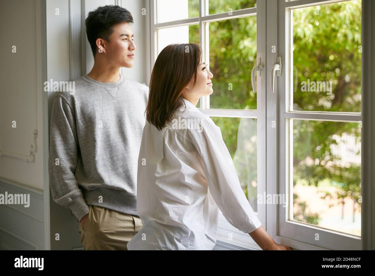 Junge asiatische paar stehen am Fenster reden während Chat Bleiben Sie zu Hause bestellen Stockfoto