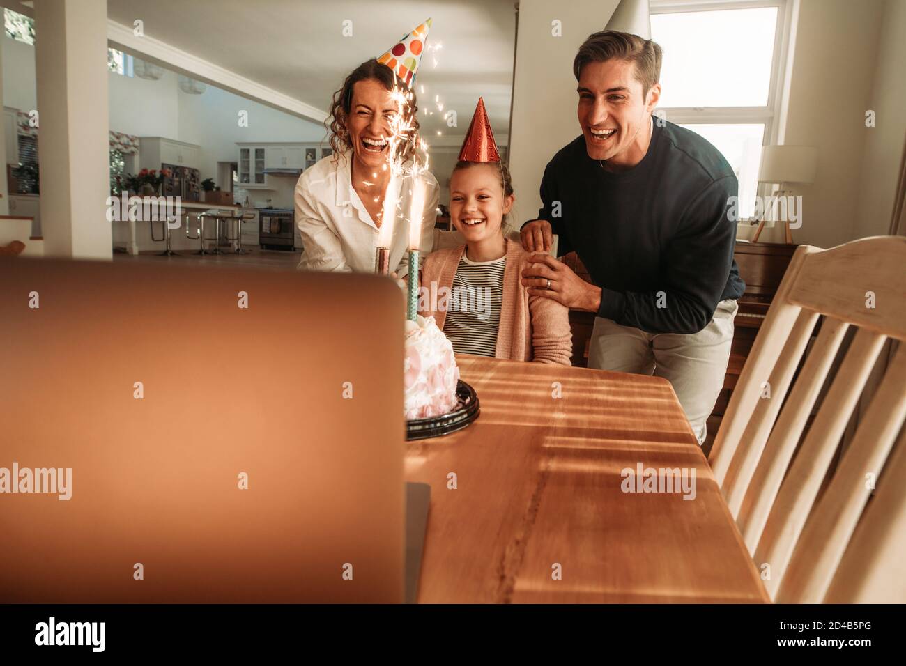 Familie feiert den Geburtstag ihrer Tochter zu Hause mit Freunden bei einem Videoanruf auf dem Laptop. Familie trägt Partyhüte mit Geburtstagskuchen auf Tisch A Stockfoto