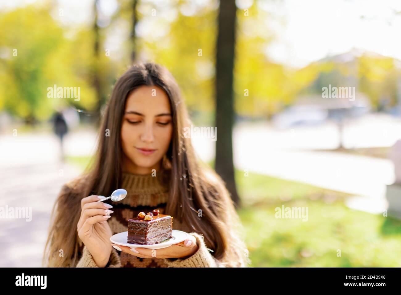 Lächelnd kaukasischen weiblichen Modell essen Phantasie Scheibe Kuchen im Freien auf dem Hintergrund des grünen Parks, genießen Sie sanfte Schokolade Dessert und Kaffee, Mädchen essen Stockfoto