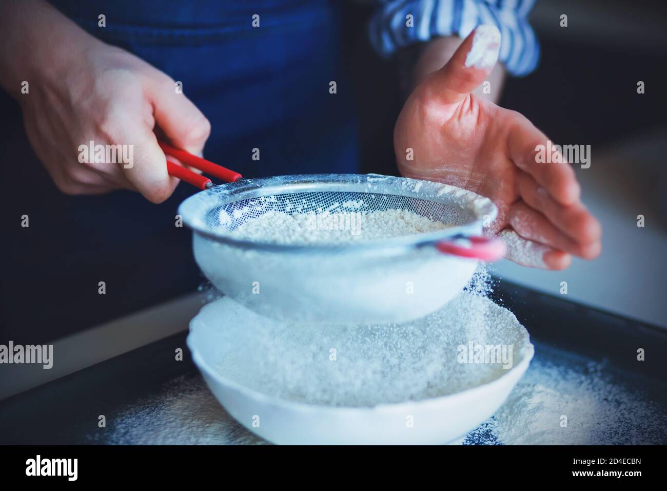 Ein Koch in einer blauen Schürze und einem gestreiften Hemd hält ein Sieb mit rotem Griff und verwendet es, um das krümelige Mehl in eine weiße Schüssel zu sieben. Hausmannskost. Stockfoto