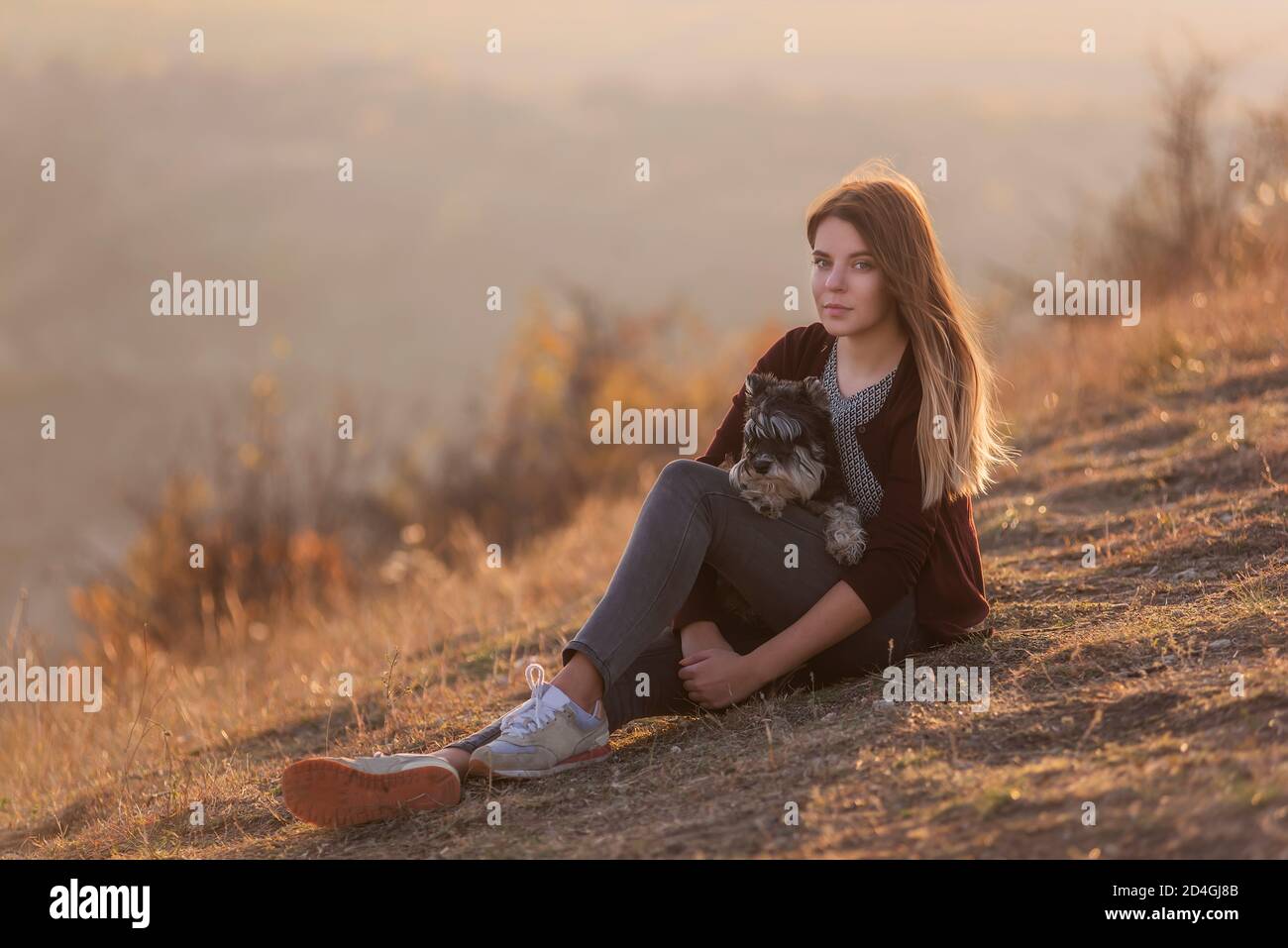 Glückliche junge Frau reist mit ihrem Haushund schwarz schnauzer. Ein schönes Mädchen umarmt ein Haustier, sitzt auf einem Berg, der Wind entwickelt ihr rotes langes Haar, Stockfoto