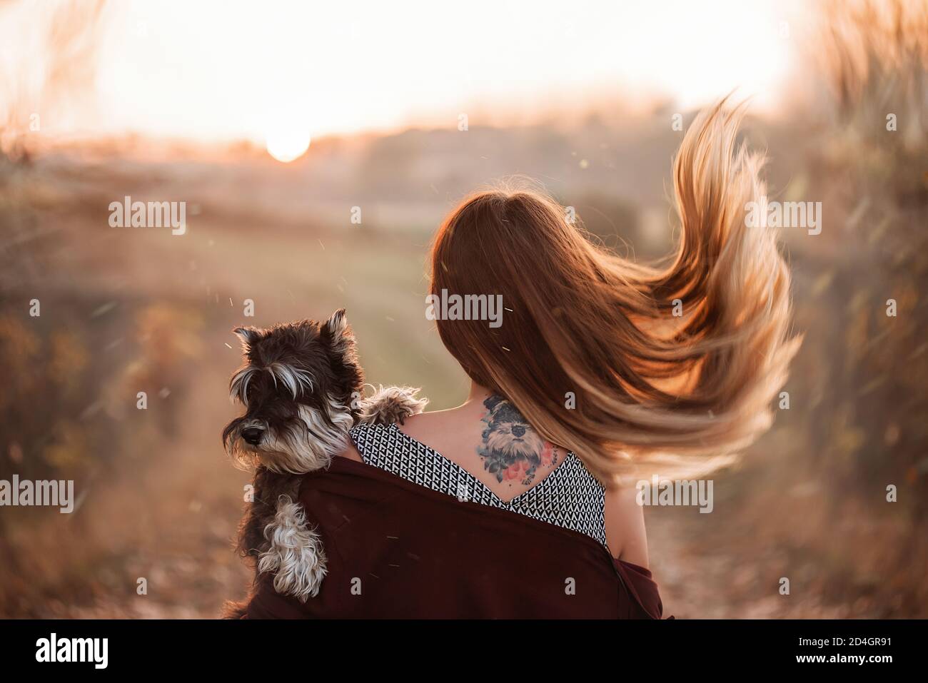 Ein blondes Mädchen mit flatternden Haaren steht auf ihrem Rücken mit dem Tattoo ihres schwarzen Schnauzer Hundes, den sie in ihrem Arm hält. PET-Hochformat. Herbst Stockfoto