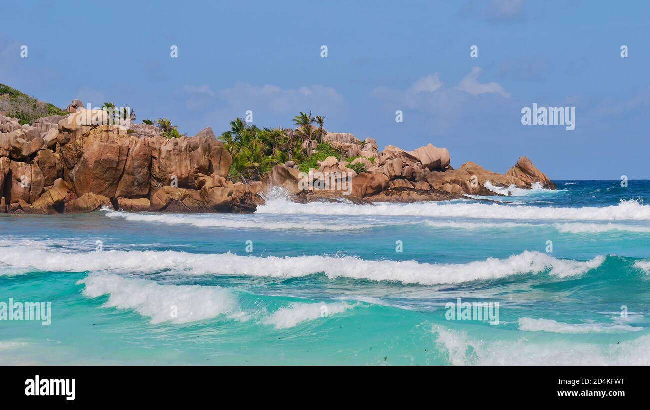 Charakteristisch schöne Granitfelsen mit Kokospalmen dazwischen und brechenden Wellen im türkisfarbenen Wasser am Strand Petite Anse, La Digue. Stockfoto