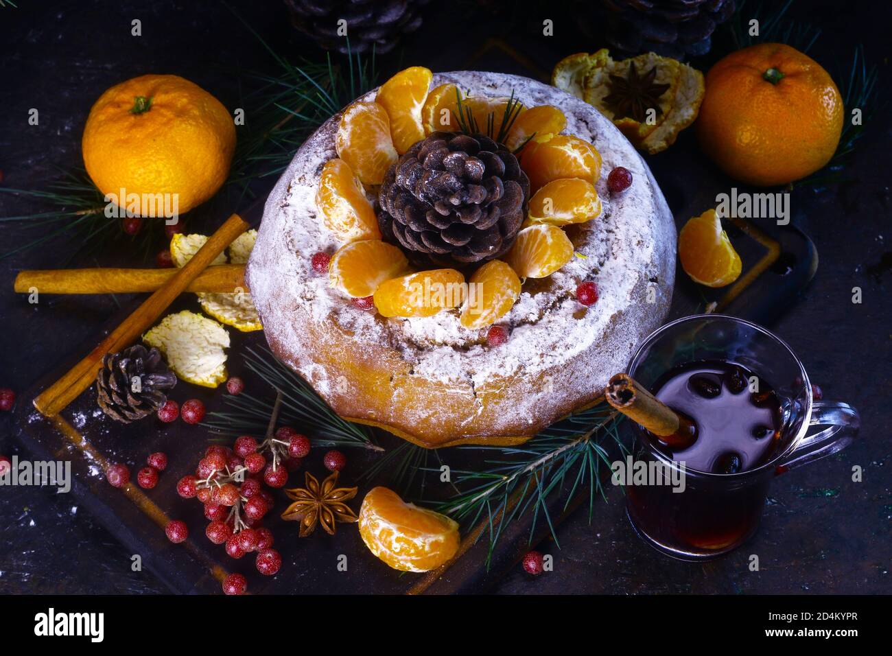 Flach liegend. Hausgemachter Kuchen mit Puderzucker und roten Beeren bedeckt. Glühwein, Tannenzweige, Tannenzapfen und Mandarinen auf dunklem Grund. Stockfoto