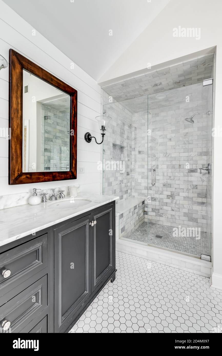 Ein luxuriöses renoviertes Badezimmer mit einem grauen Waschtisch, rustikalem Holzspiegel und einer Duschkabine mit Glastür und benutzerdefinierten Marmorfliesen. Stockfoto