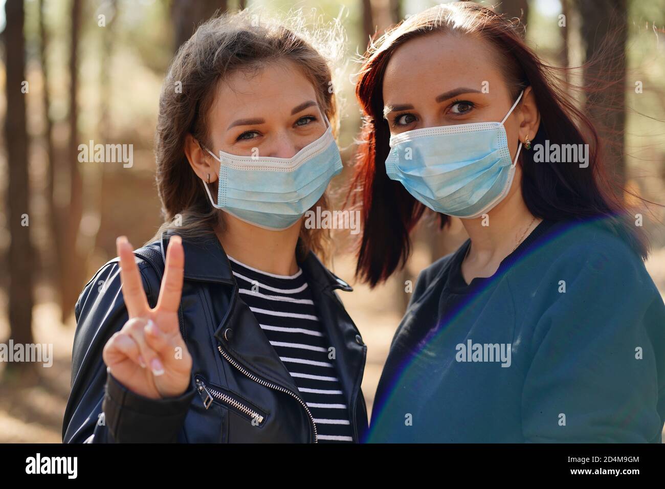 Zwei junge Frauen in medizinischen Masken betrachten die Kamera in Holz. Nahaufnahme von Frauen schützen sich vor Krankheiten auf dem Spaziergang. Konzept der Bedrohung von Stockfoto