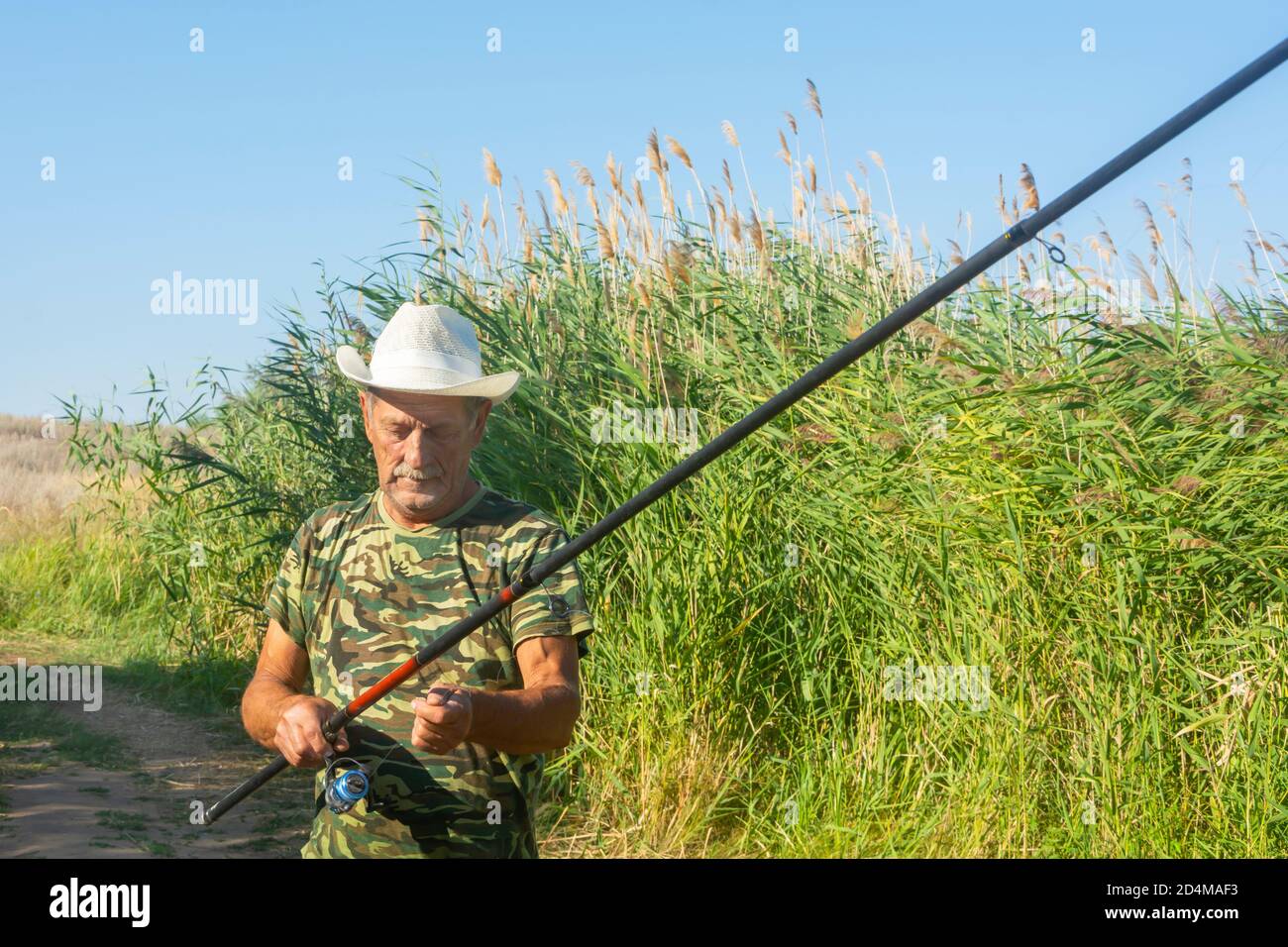 Senior Fischer im Hut mit einem Angelrute bereitet sich auf das Fischen am Ufer mit Schilf. Ein älterer Rentner ruht in der Natur. Weichfokus. Stockfoto