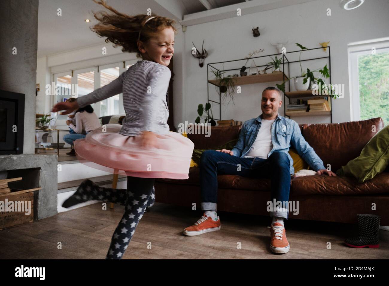 Junge Tochter tanzt herum, während Vater glücklich auf dem Sofa zuschaut Des Wohnzimmers Stockfoto