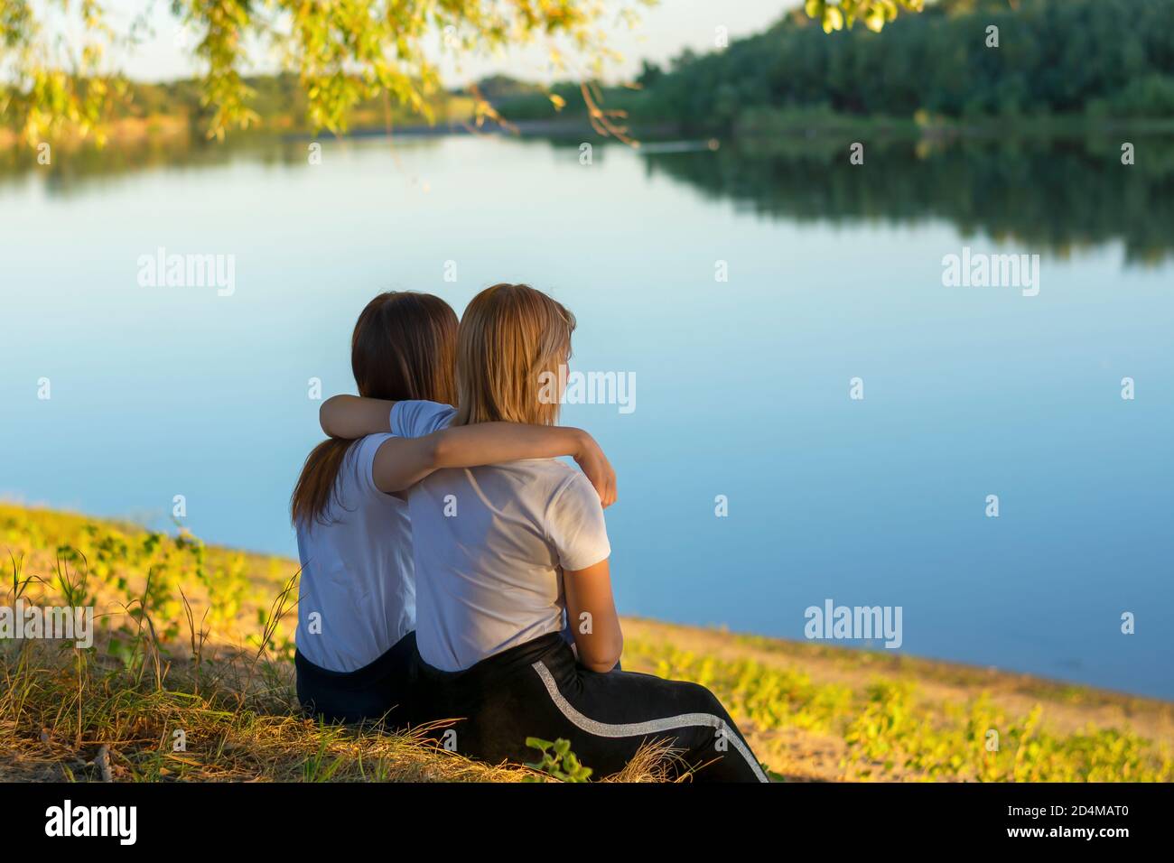 Zwei junge Frauen sitzen im Herbst auf dem Gras in der Nähe des Flusses. Freundinnen umarmen, Rückansicht. Stockfoto
