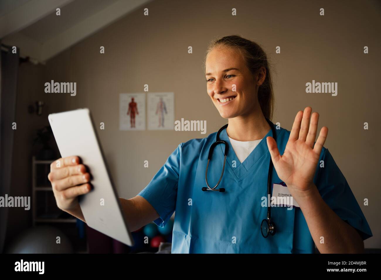 Schöne junge kaukasische Ärztin auf Videoanruf aus der Praxis zu Hause, lächelnd und winkend an Patienten Stockfoto